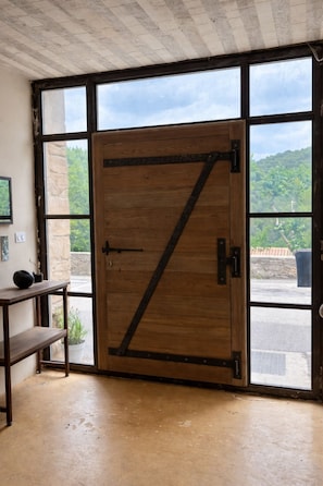 Eine Holztür mit diagonalem Metallstab, ein kleiner Tisch mit einer Vase und Blick auf Grün durch große Fenster.