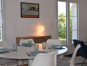 Ein Essbereich mit einem runden Tisch, weißen Stühlen und einer Bank. An der Wand hängt ein gerahmtes Bild, es gibt eine Lampe und große Fenster mit Blick auf Grünflächen.
