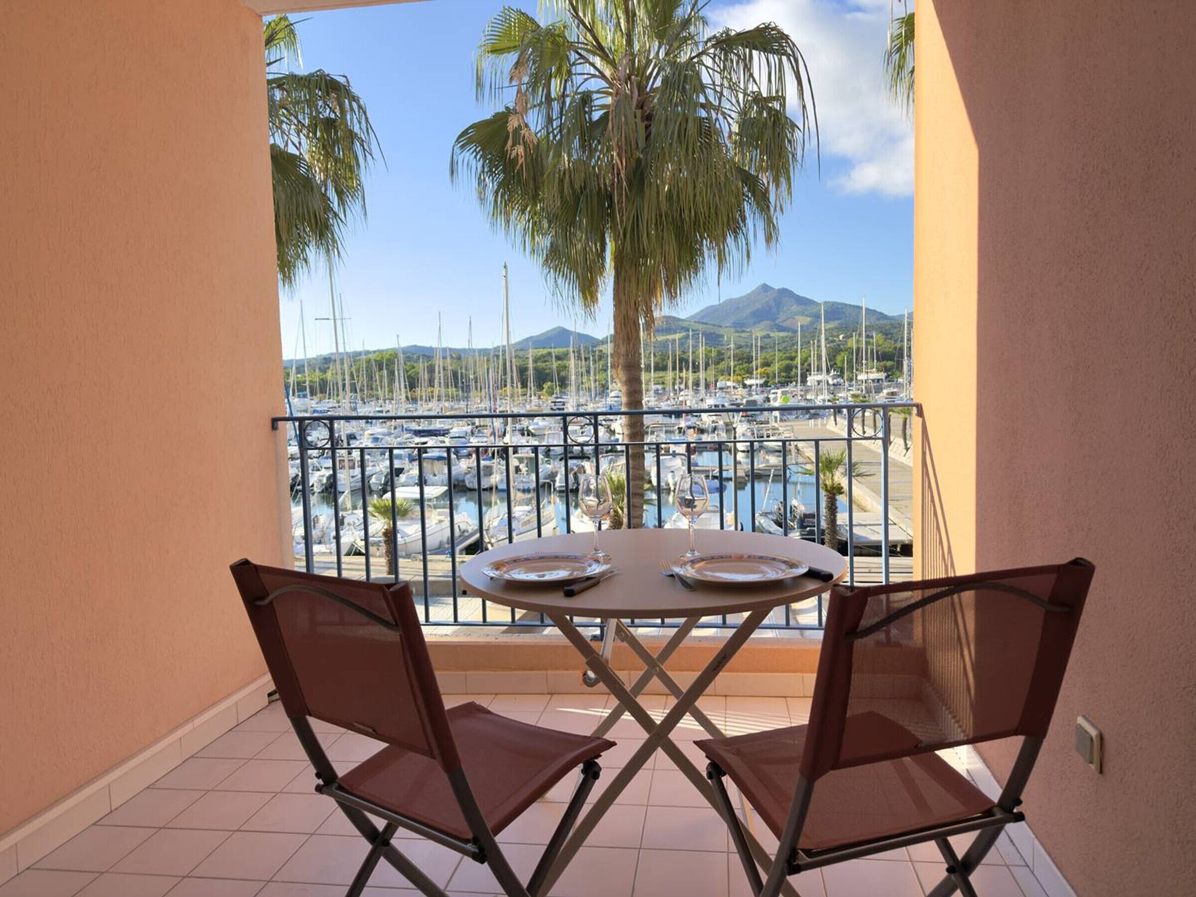 Ein Balkon mit Tisch und Stühlen, mit Blick auf einen Yachthafen mit Booten und einem Berg im Hintergrund.