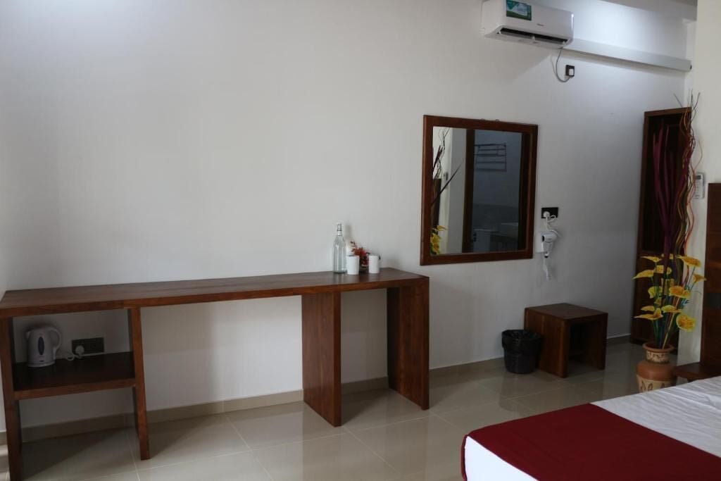 A hotel room with a wooden desk, a bed, a mirror, an air conditioner, a small stool, and a trash bin.