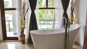 Um banheiro moderno com uma banheira independente, uma janela grande com cortinas, e vista para uma piscina e palmeiras.