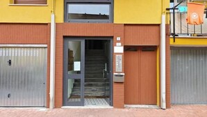 Una entrada de edificio con puerta de cristal, escalera y un pequeño espacio exterior con barandilla.