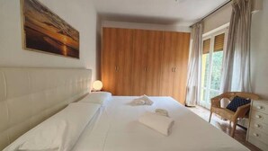 Una cama bien hecha con edredón blanco y almohadones, un armario de madera, una silla de mimbre con cojín y un cuadro en la pared.