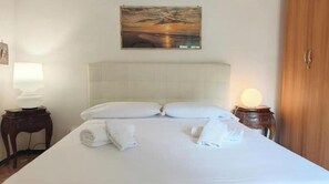 Una cama bien hecha con almohadas y toallas, un cuadro en la pared, una lámpara en una mesita de noche y una puerta de madera.