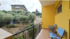 Un balcón con mesa y sillas, una pared amarilla y vistas a un edificio y vegetación.