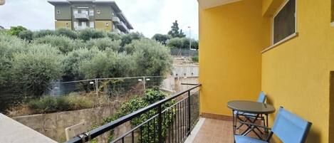 A balcony with a table and chairs, a yellow wall, and a view of a building and greenery.