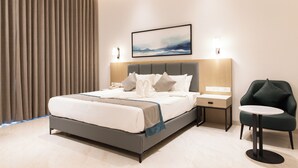 A modern hotel room with a large bed, bedside tables, a small round table, a chair, and a painting on the wall. - Lore Resorts and conventions (Maheshwaram)