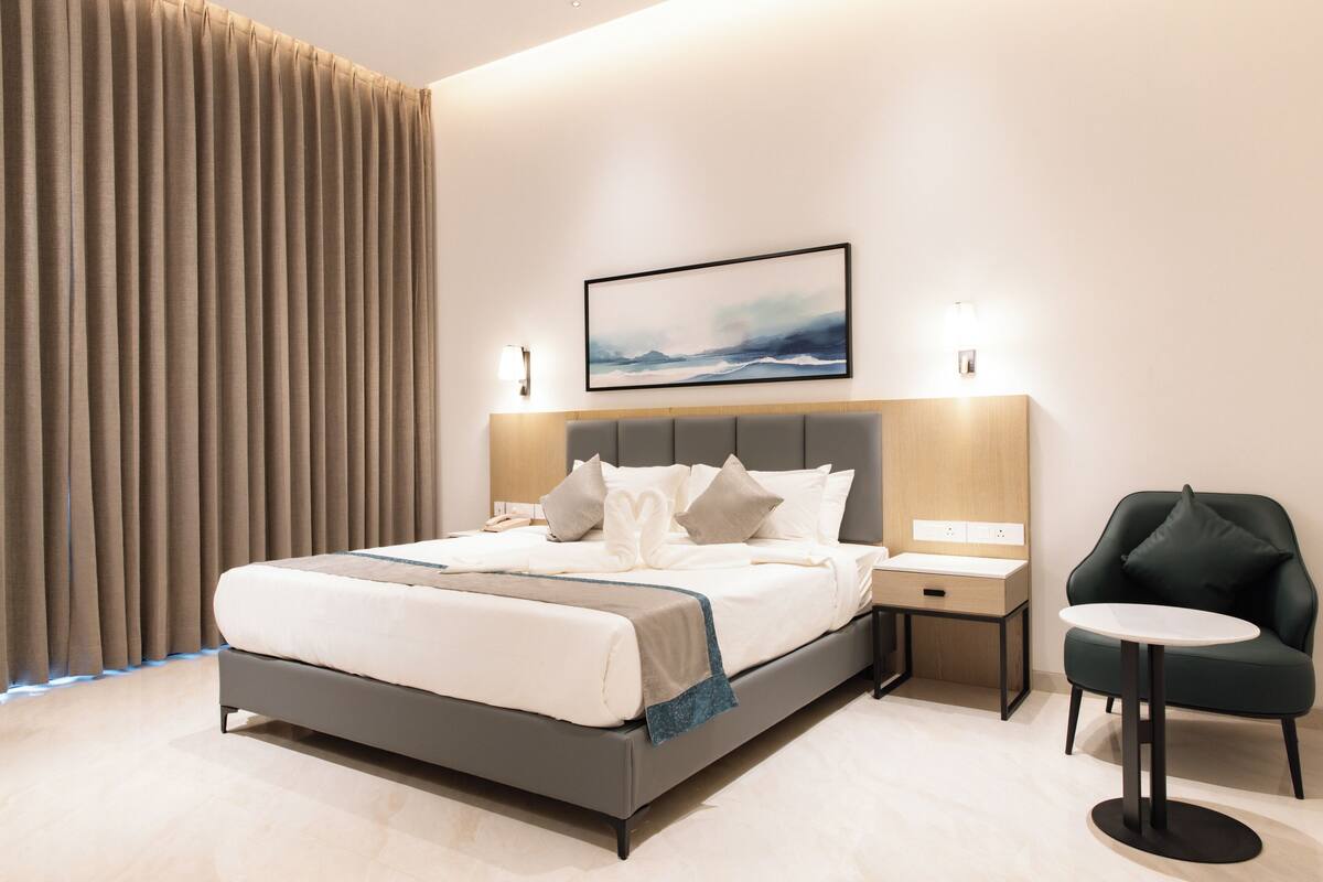 A modern hotel room with a large bed, bedside tables, a small round table, a chair, and a painting on the wall.