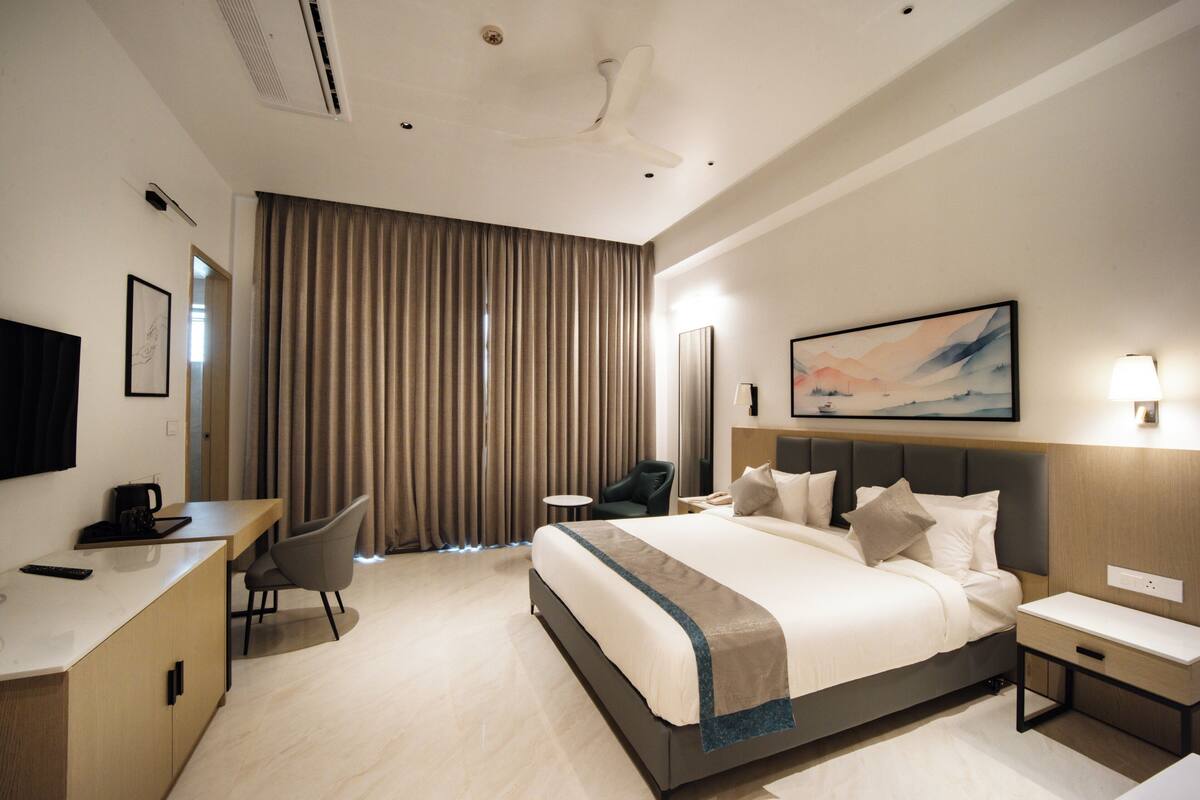 A hotel room with a bed, desk, chair, bedside table, and a painting on the wall.