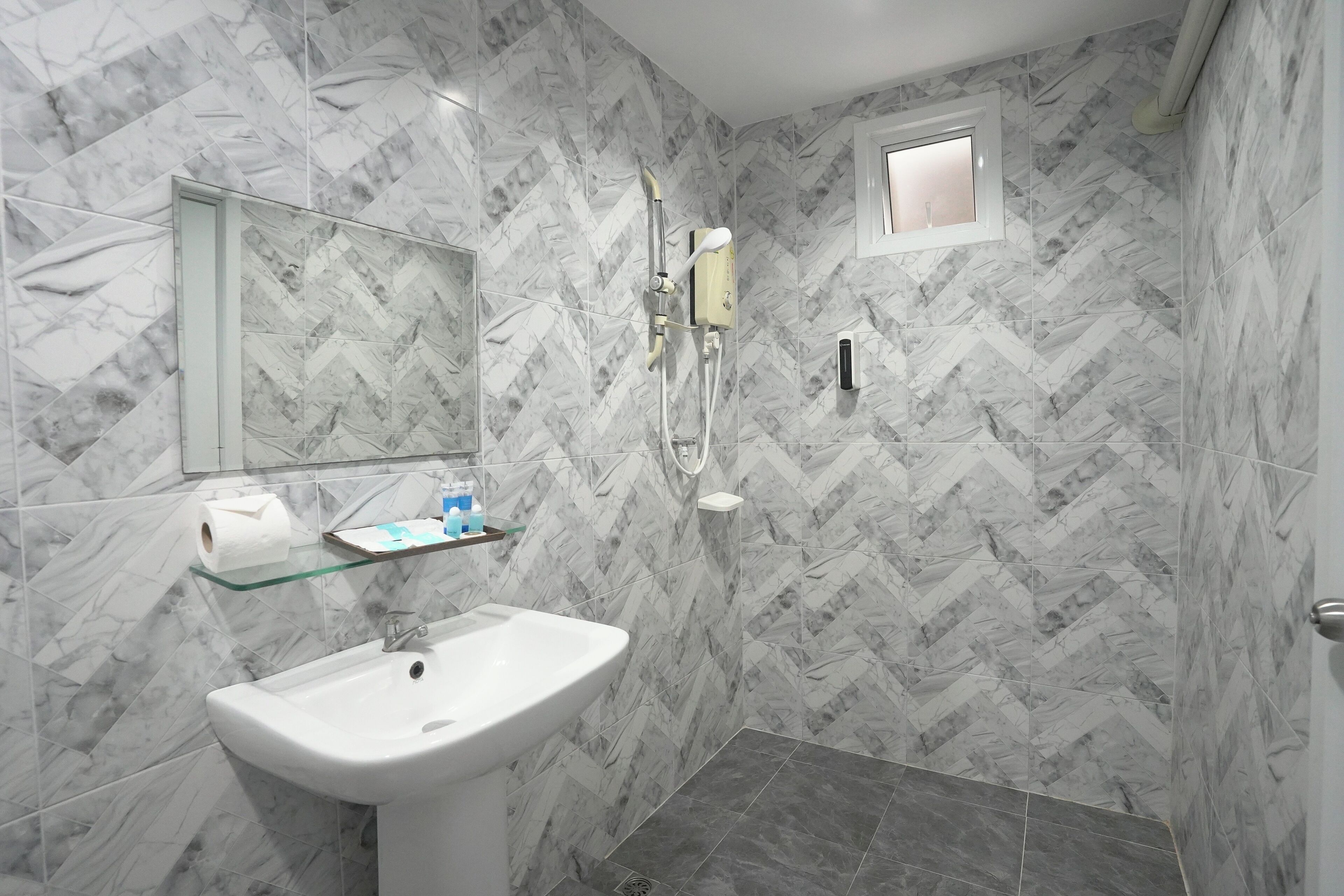 A bathroom with a marble herringbone pattern, a white sink, a glass shelf with toiletries, and a shower with a handheld showerhead.