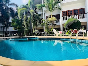 Uma piscina com cerca branca, rodeada por palmeiras e um edifício com varandas.