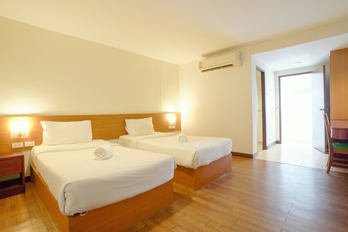 A hotel room with two beds, wooden flooring, a wall-mounted air conditioner, and a door leading to another room.