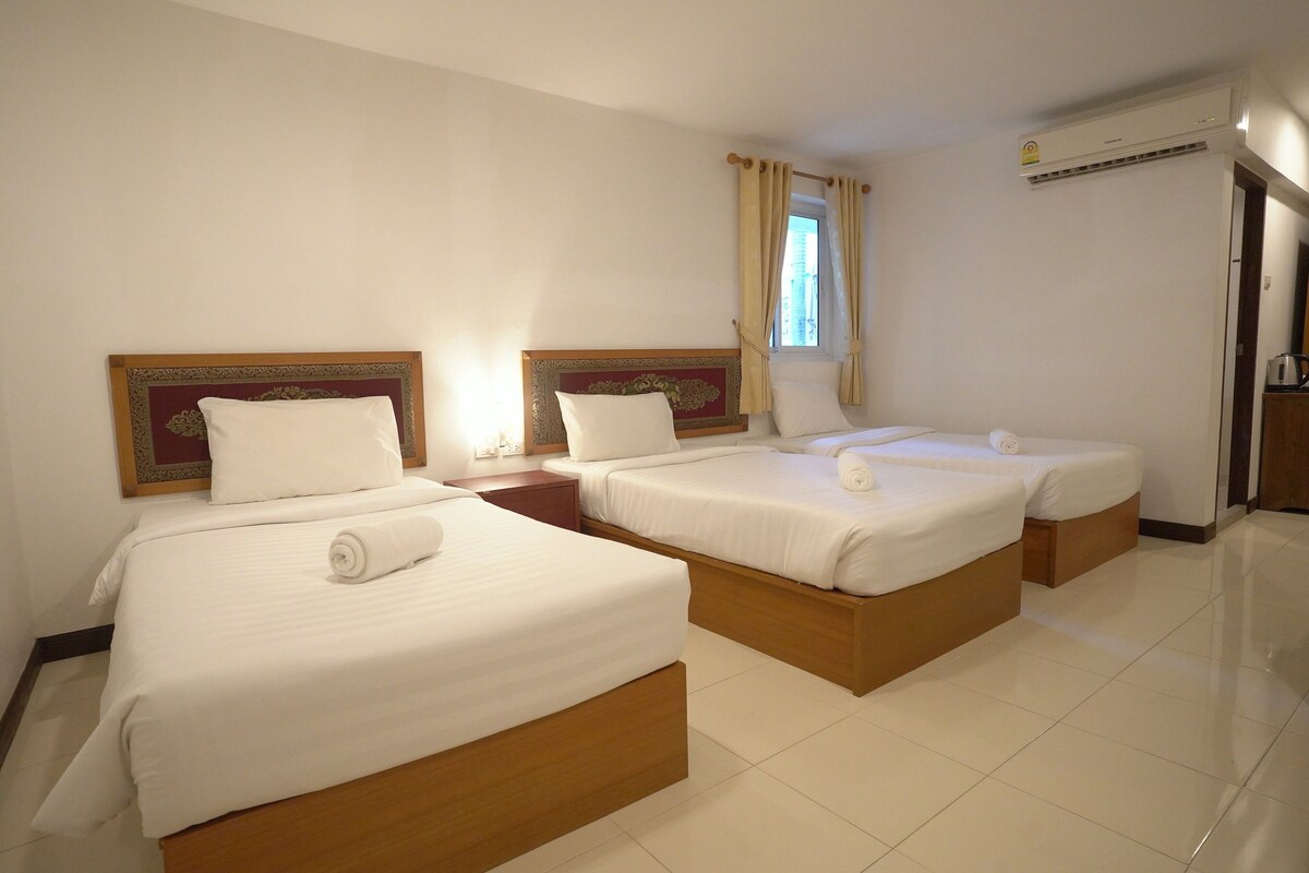 A hotel room with three single beds, a nightstand, a window with curtains, and an air conditioning unit.