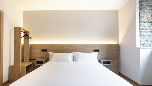 Una habitación de hotel moderna con una cama grande, cabecera de madera, mesitas de noche y una lámpara fijada a la pared.