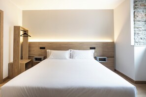 Une chambre d’hôtel moderne avec un grand lit, une tête de lit en bois, des tables de chevet et un luminaire fixé au mur.