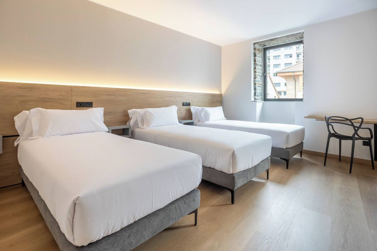 Une chambre d’hôtel avec trois lits, un bureau et une fenêtre donnant sur des immeubles.