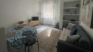 Ein Wohnzimmer mit Deckenventilator, einer Couch, einem Fernseher auf einem Schrank und einem Esstisch mit gemustertem Tischläufer.