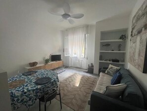 A living room with a ceiling fan, a sofa, a TV on a stand, and a dining table with a patterned tablecloth. - Hostdomus - Sun House (Finale Ligure)