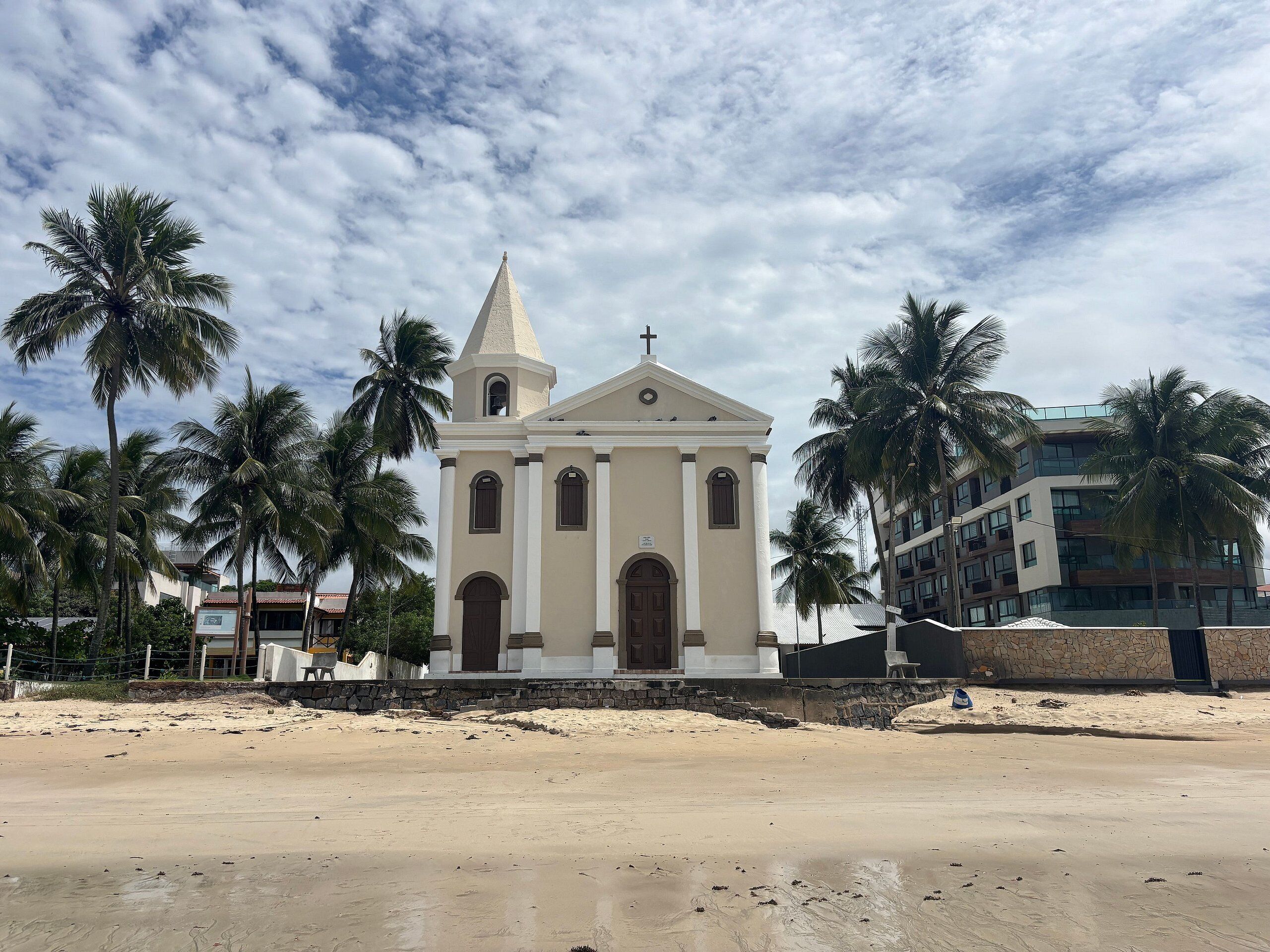 A church with a bell tower, surrounded by palm trees and a sandy beach.