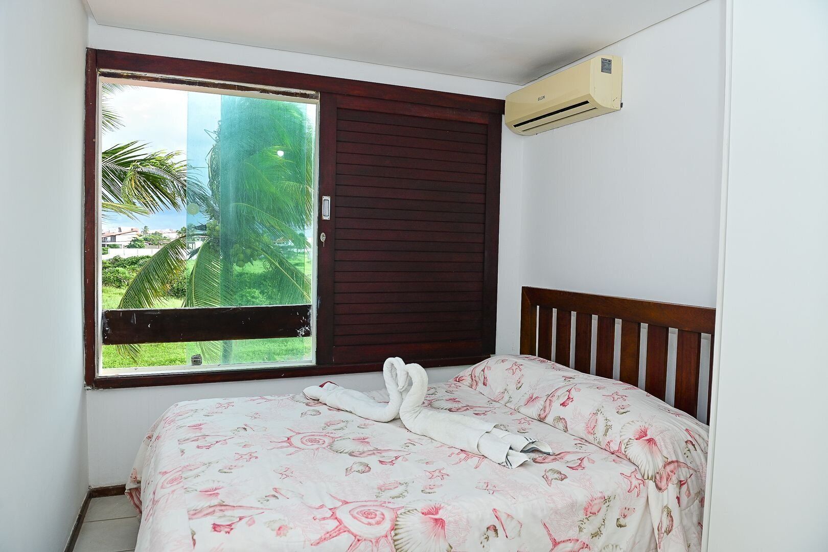 A room with a bed, a window with a view of greenery, and an air conditioning unit.