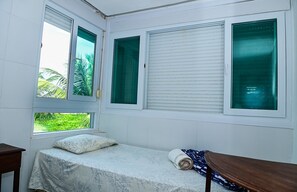Una habitación pequeña con cama, dos ventanas con persianas y vistas al exterior con vegetación.
