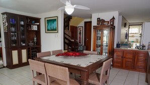 A dining area with a wooden table, wicker chairs, a ceiling fan, and a kitchen in the background. - Piratas do Caribe - Casa das Conchas (Tamandaré)