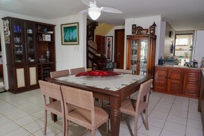 Un comedor con una mesa de madera, sillas de mimbre, un ventilador de techo y una cocina al fondo.