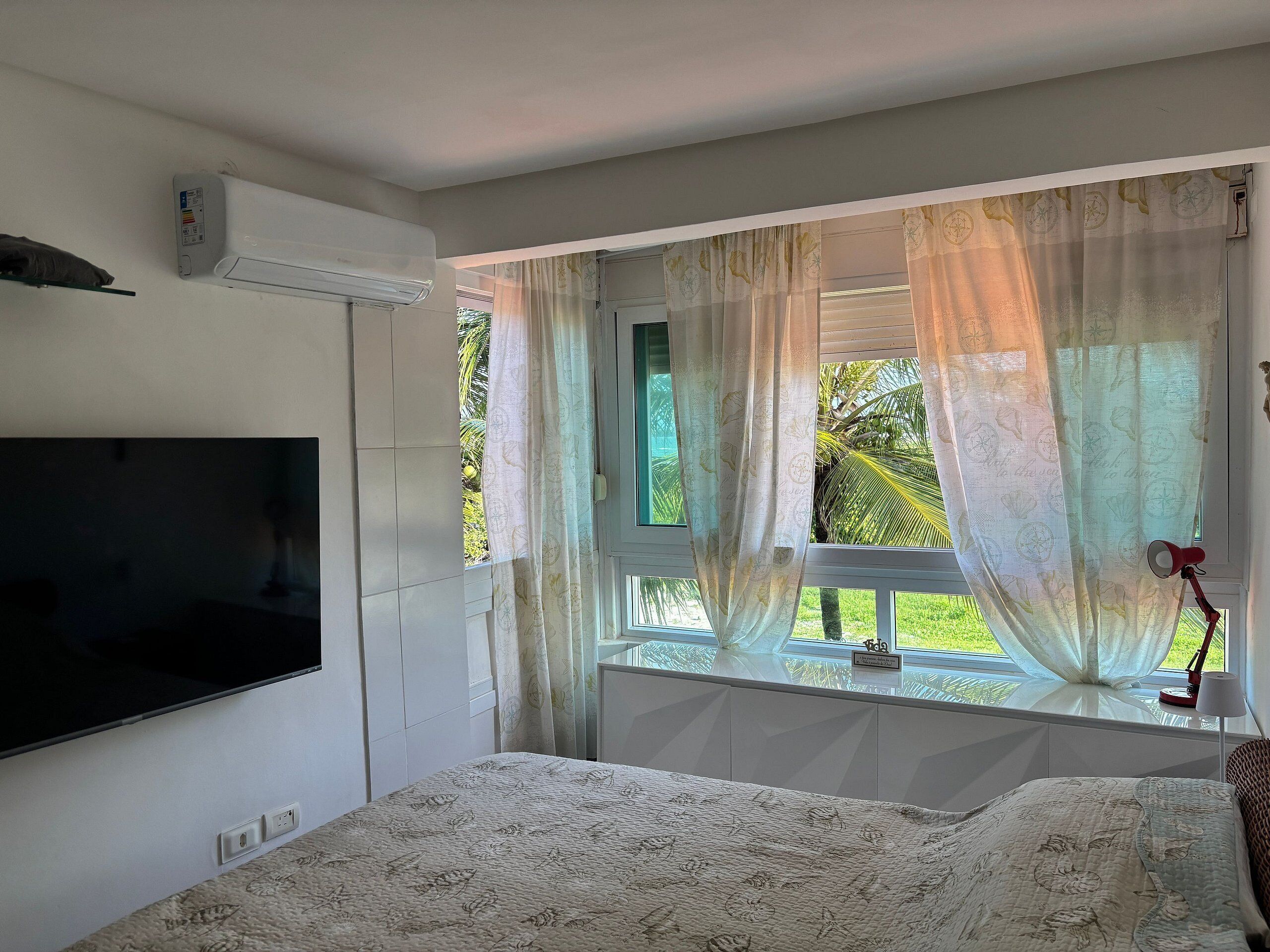 A bedroom with a bed, a TV mounted on the wall, a window with sheer curtains, and a view of greenery outside.