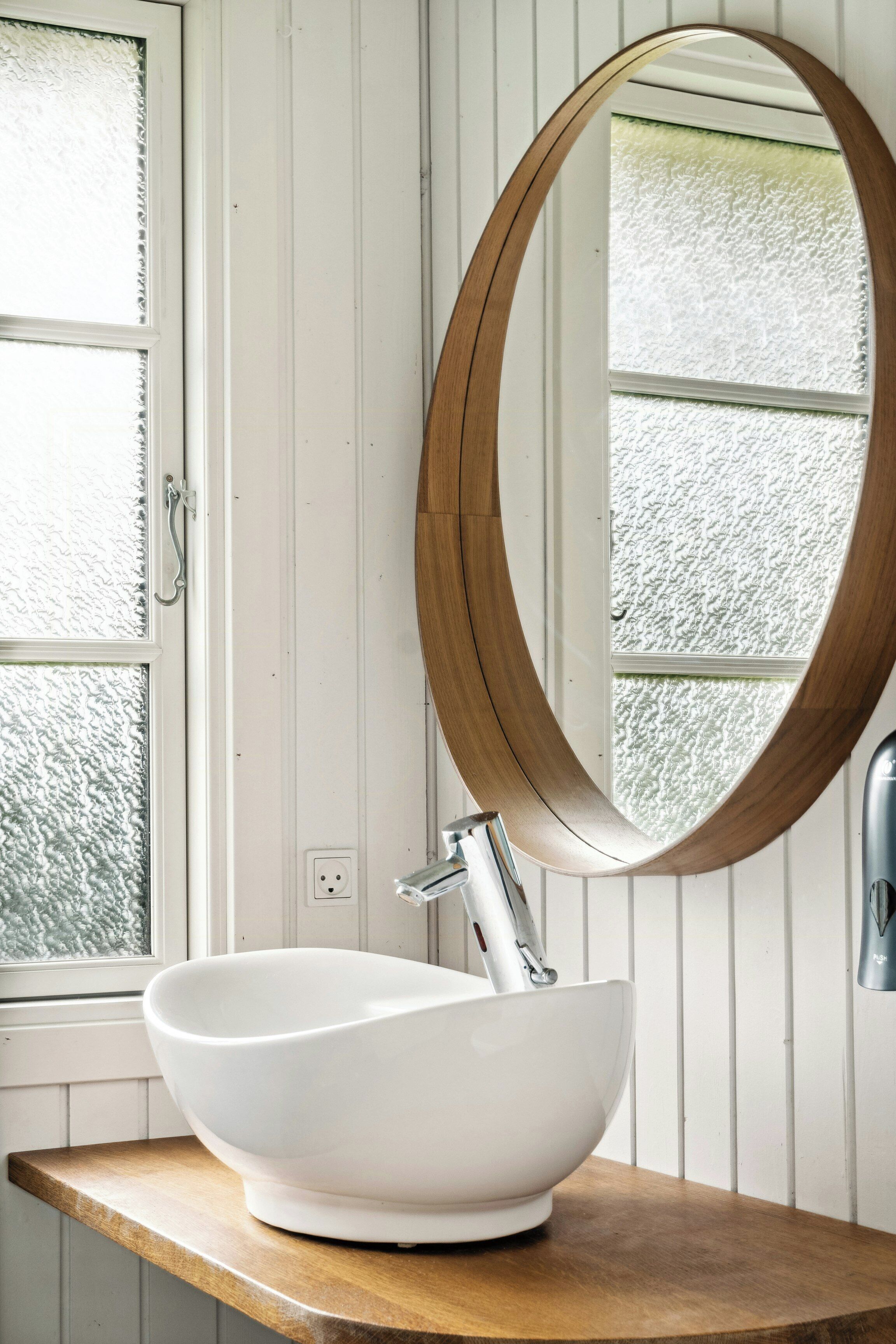 A white ceramic sink with a modern faucet, a wooden-framed mirror, and a window with frosted glass.