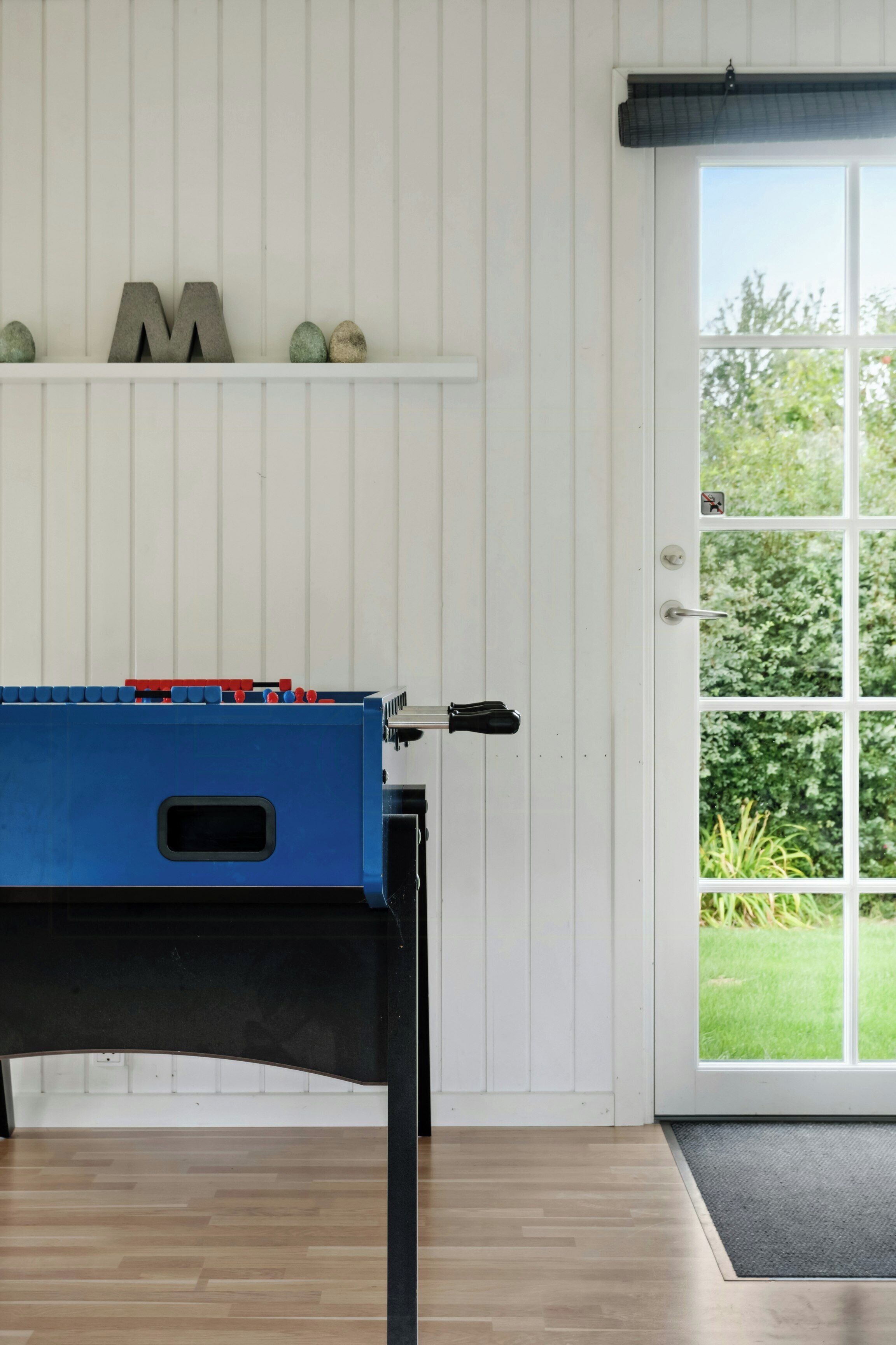 Une pièce avec un plancher en bois, une table bleue recouverte d’une nappe noire, une porte blanche et une fenêtre donnant sur un paysage verdoyant.