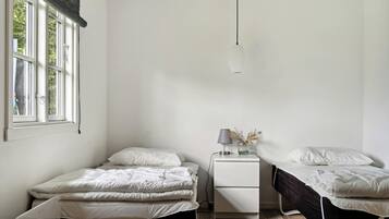 Ein Schlafzimmer mit zwei Einzelbetten, einem Nachttisch mit Lampe und einem Fenster mit Blick auf Bäume.