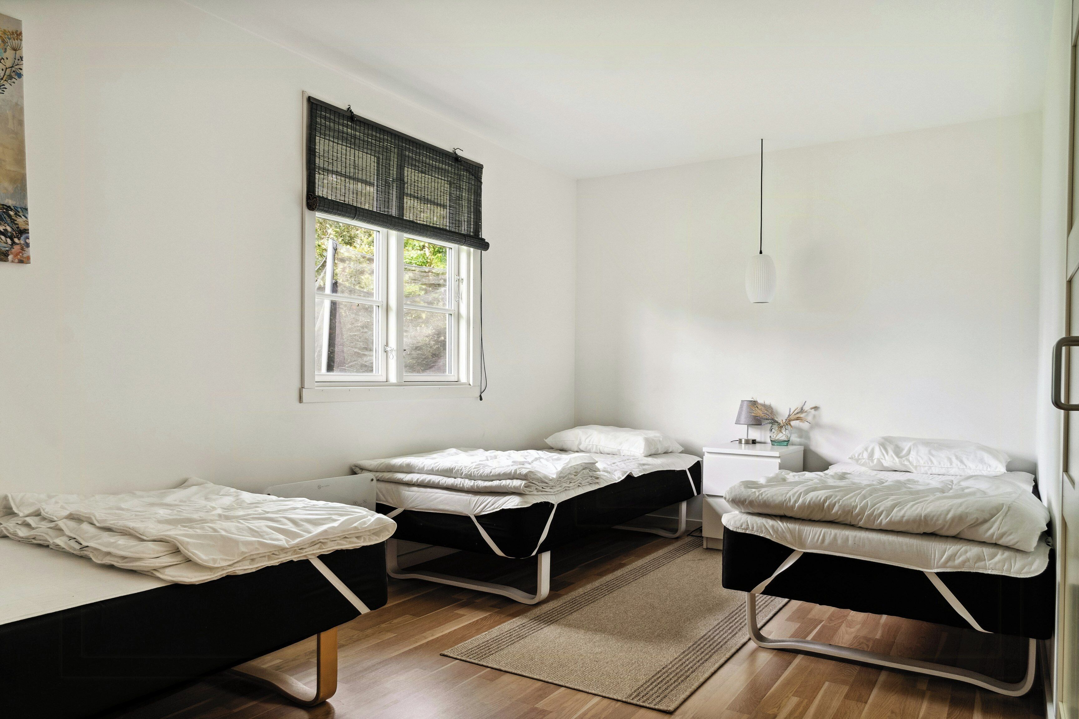 Ein Zimmer mit drei Einzelbetten, einem Fenster mit schwarzem Rollo, einem Nachttisch mit Lampe und einer Deckenleuchte.