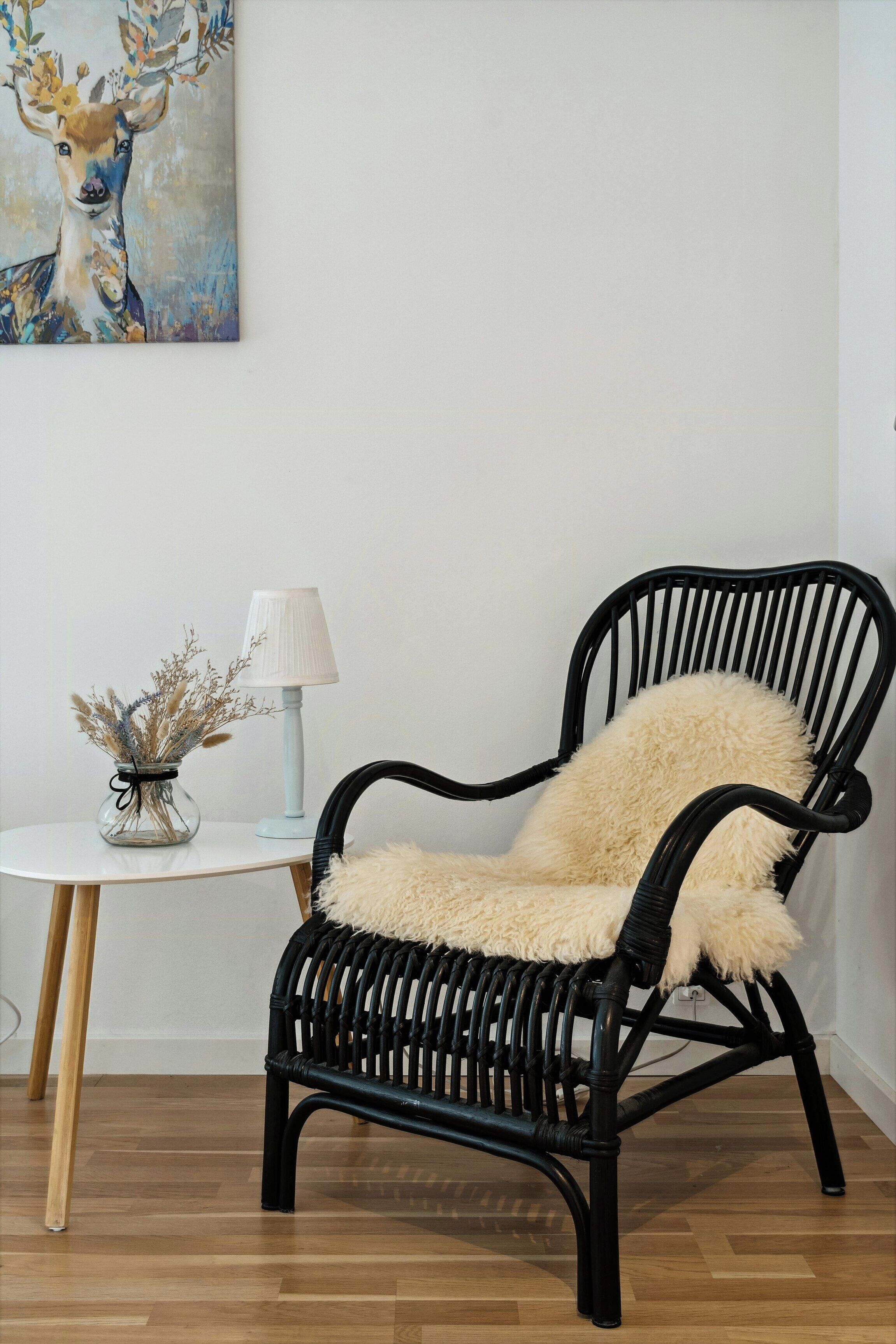 A black wicker chair with a fluffy beige cushion, a white table with a lamp and a vase of dried flowers, and a painting of a deer on the wall.