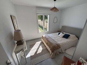 A bedroom with a bed, a desk, a lamp, a window, and a hanging light fixture. - Maison médis 2 bedrooms, sleeps 6 (Médis)