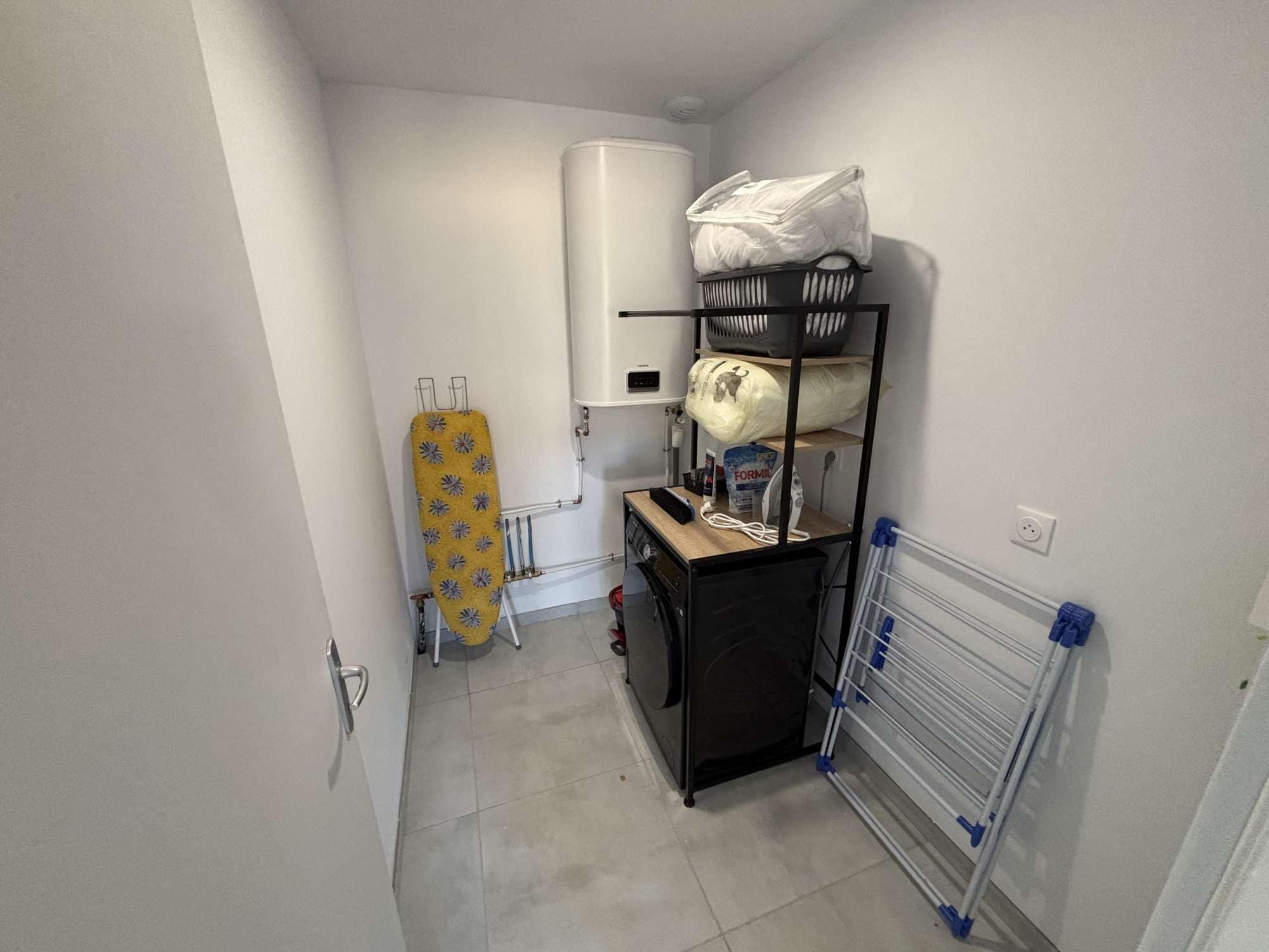 A compact laundry room with a washing machine, ironing board, and drying rack.