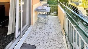 Varanda com mesa e cadeiras, porta de vidro deslizante e vista para um edifício e árvores.