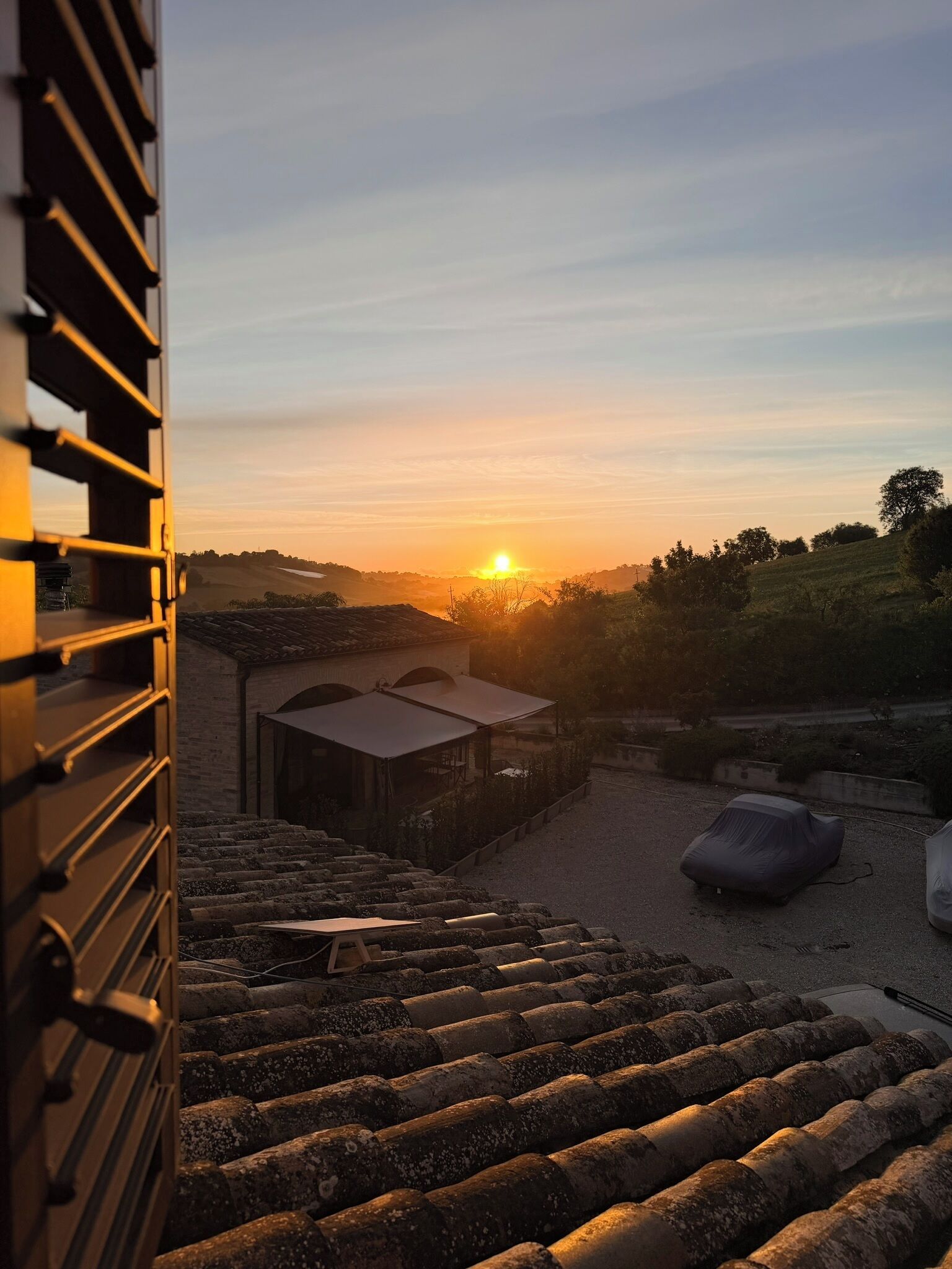 A sunset view from a building with a staircase leading to a covered area, a car, and a rural landscape.