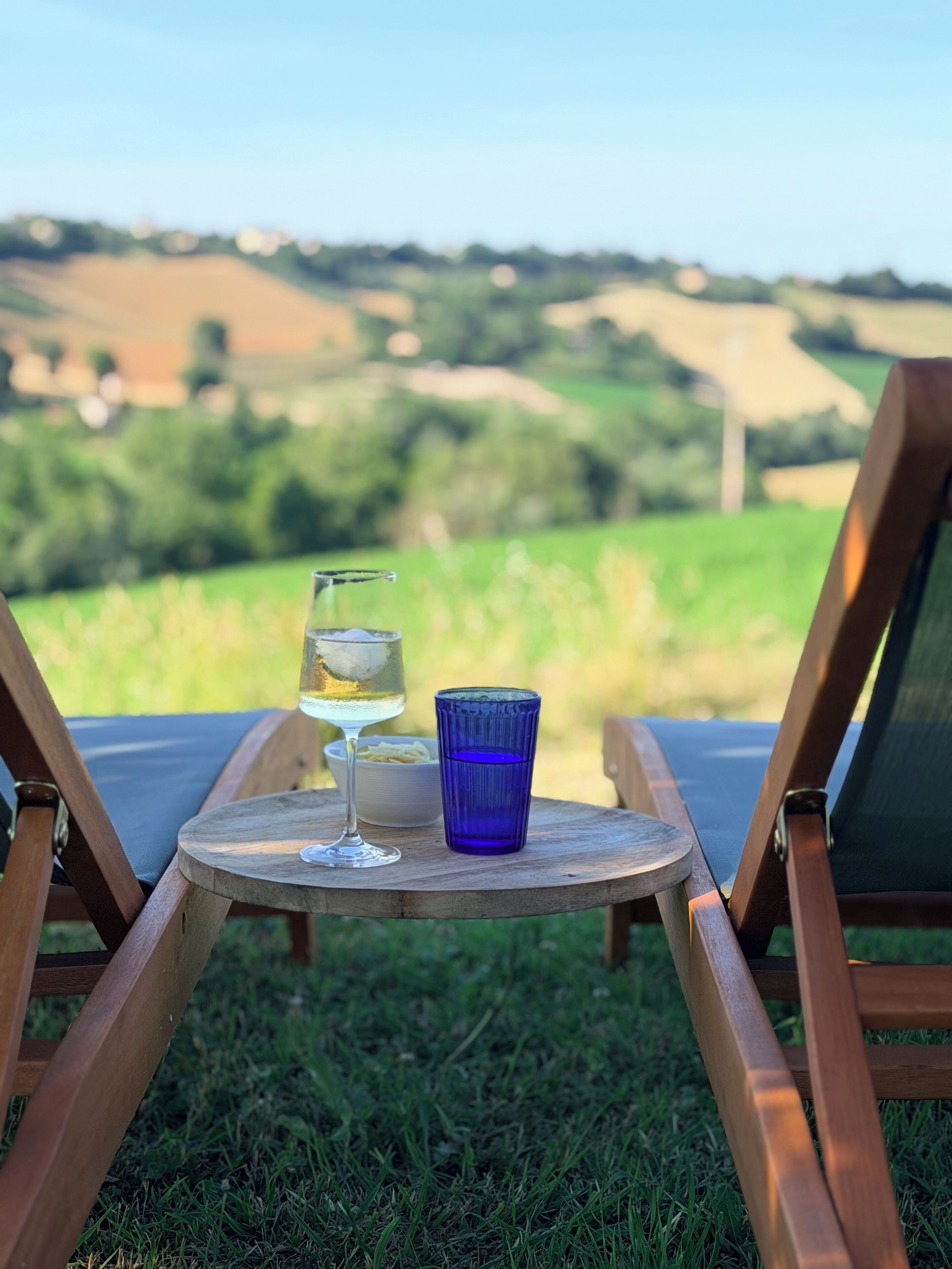 A wooden table with a glass of white wine and a blue glass on a grassy area with a view of green fields.