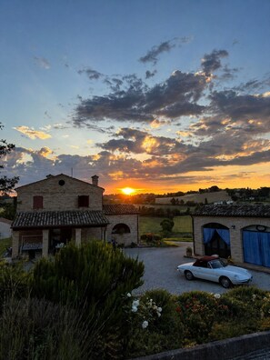 A rural landscape with a sunset, a stone building, and a white car parked in front.
