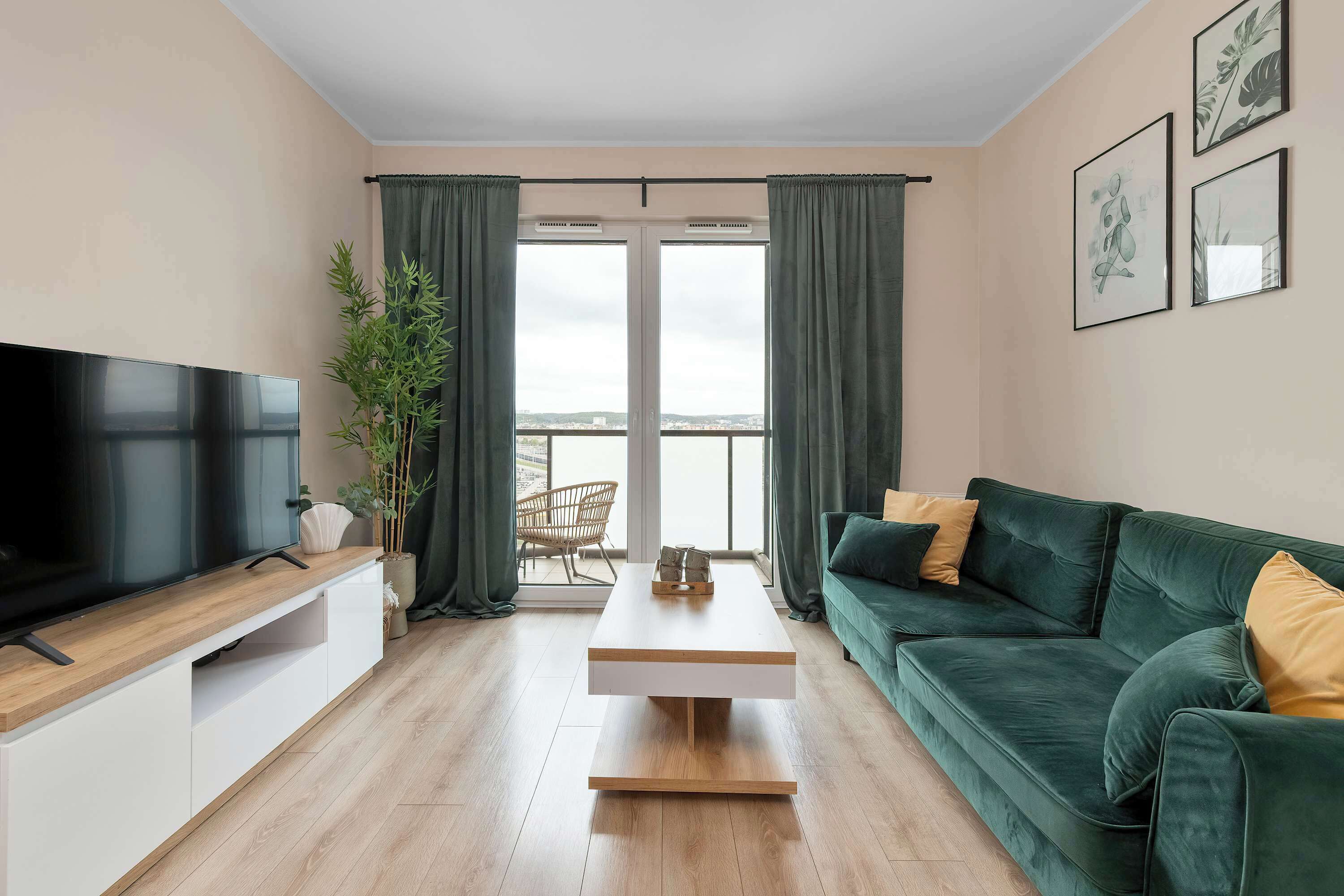 A modern living room with a flat-screen TV, a wooden coffee table, a green sofa, and a balcony with curtains.