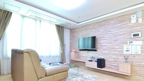 A modern living room with a leather sofa, a flat-screen TV mounted on a wood-patterned wall, a glass coffee table, and a small side table with a speaker. - GeojeResidenceHotel (Geoje)
