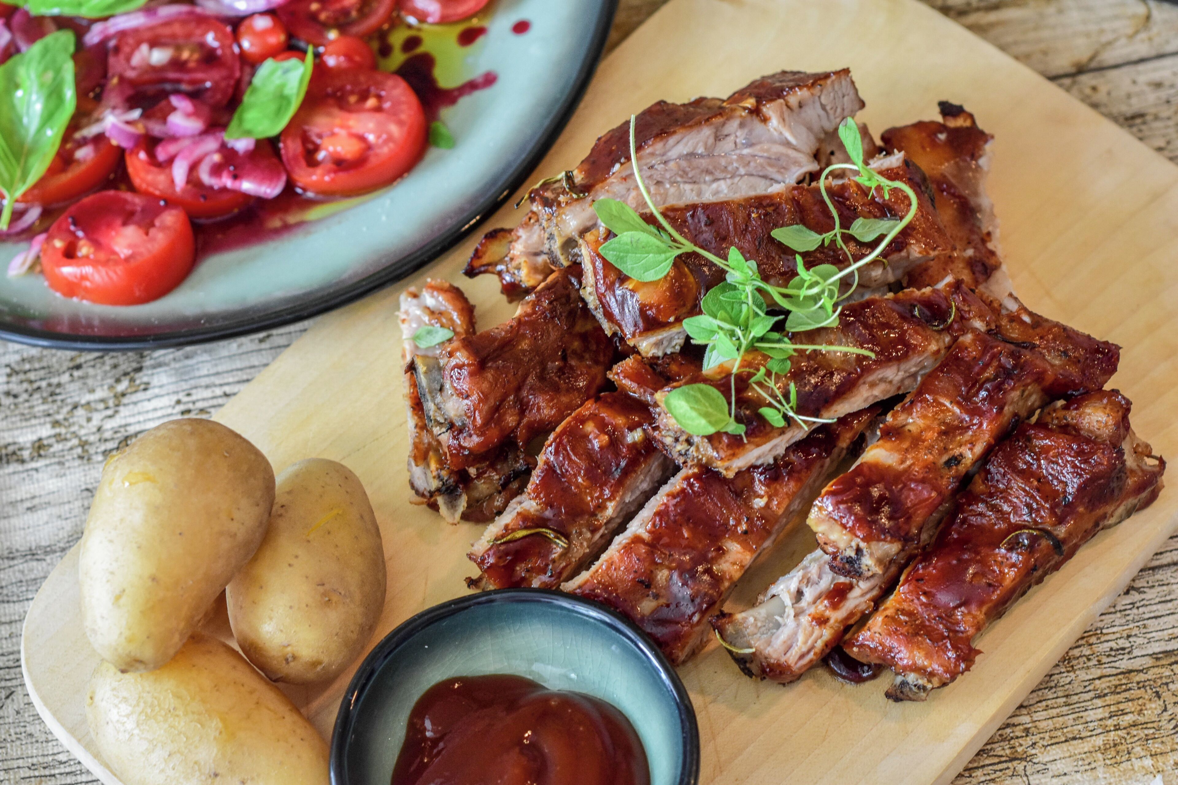A wooden cutting board with sliced meat, potatoes, and a bowl of sauce.