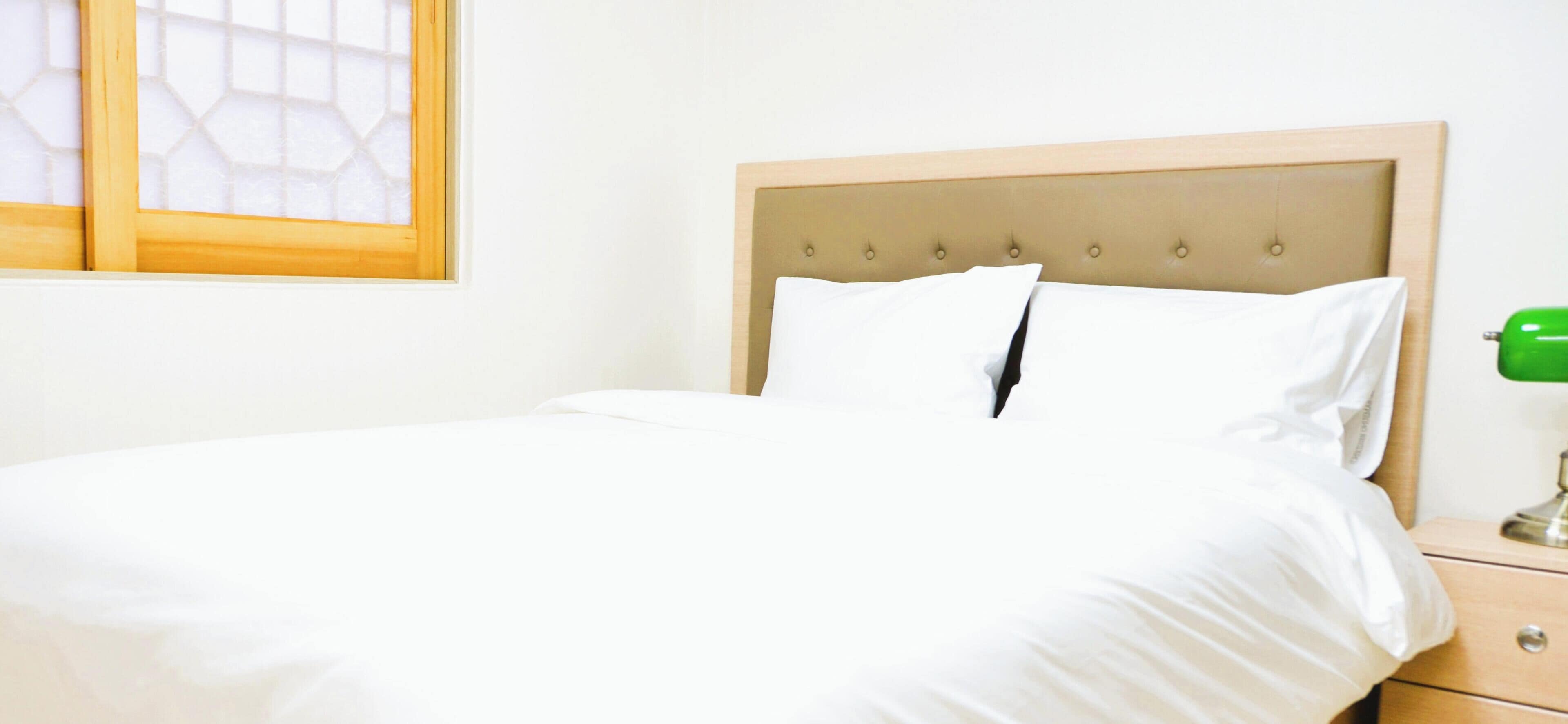 A neatly made bed with a wooden headboard, a bedside table with a green lamp, and a window with a geometric pattern.