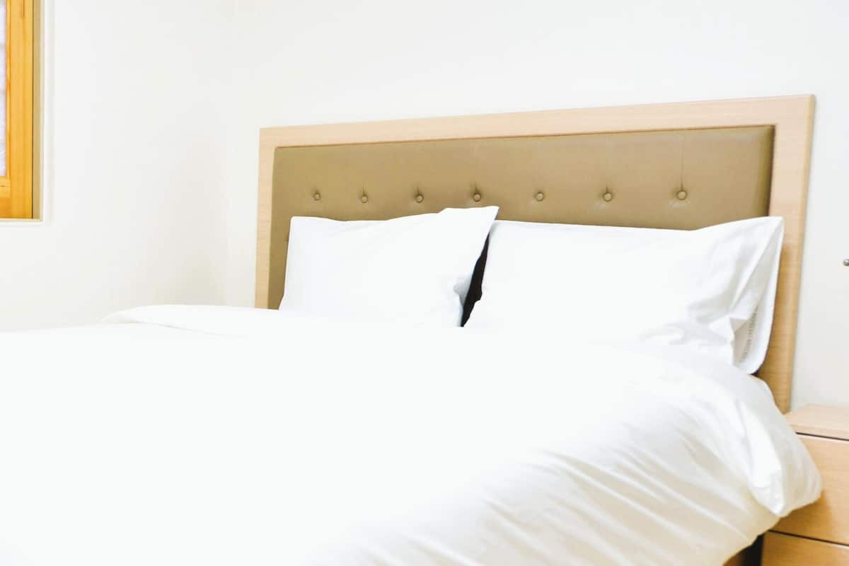 Una cama bien hecha con cabecera de madera, una mesita de noche con una lámpara verde y una ventana con diseño geométrico.