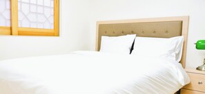 Una cama bien hecha con cabecera de madera, una mesita de noche con una lámpara verde y una ventana con diseño geométrico.