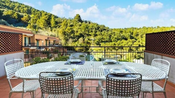 A terrace with a table and chairs, a view of a forested hillside, and a building in the background. - Hostdomus - Olive Grove House (Laigueglia)