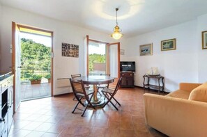 Een zithoek met een ronde tafel en stoelen, een bank, een televisie en een balkon met uitzicht op groen.