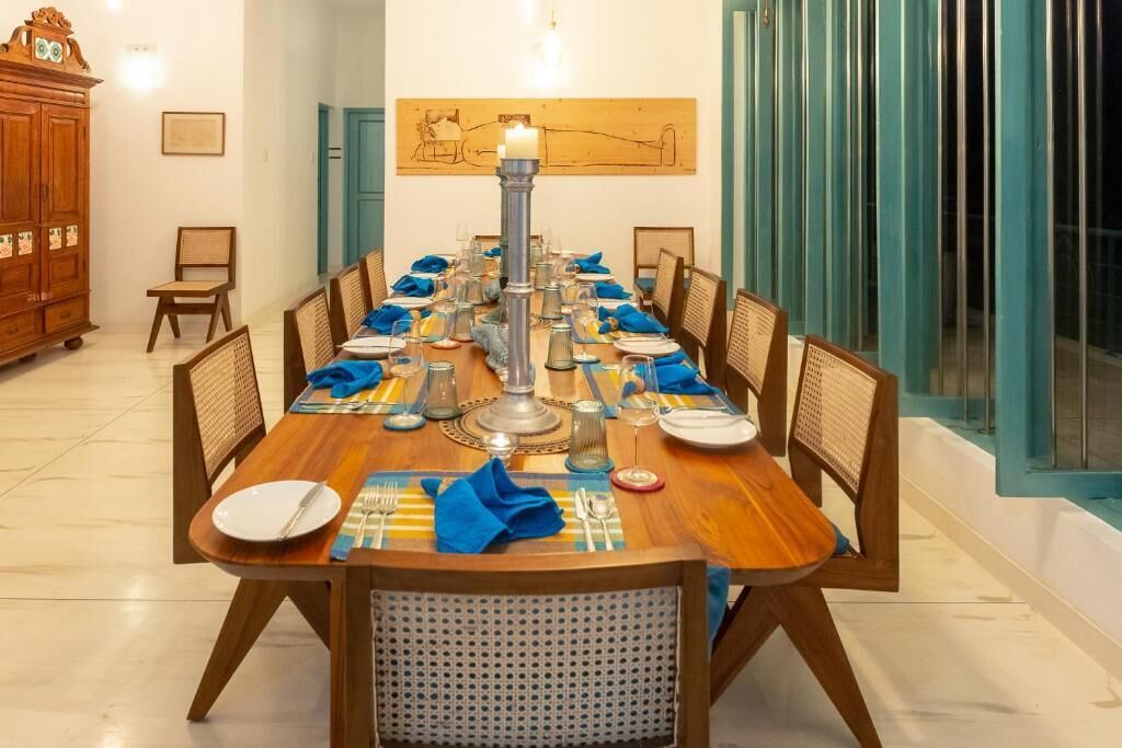 A dining area with a long wooden table set for a meal, surrounded by chairs, with a large painting on the wall and a cabinet to the left.