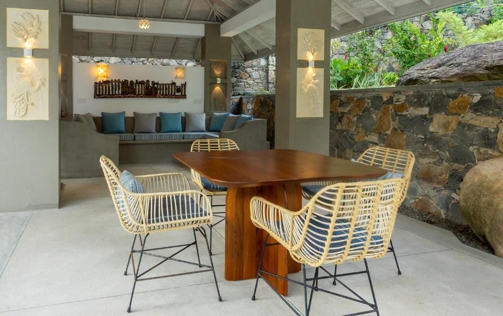 Une terrasse extérieure avec une table en bois et des chaises en osier, un mur en pierre et un coin salon agrémenté de coussins et d’œuvres d’art mural.