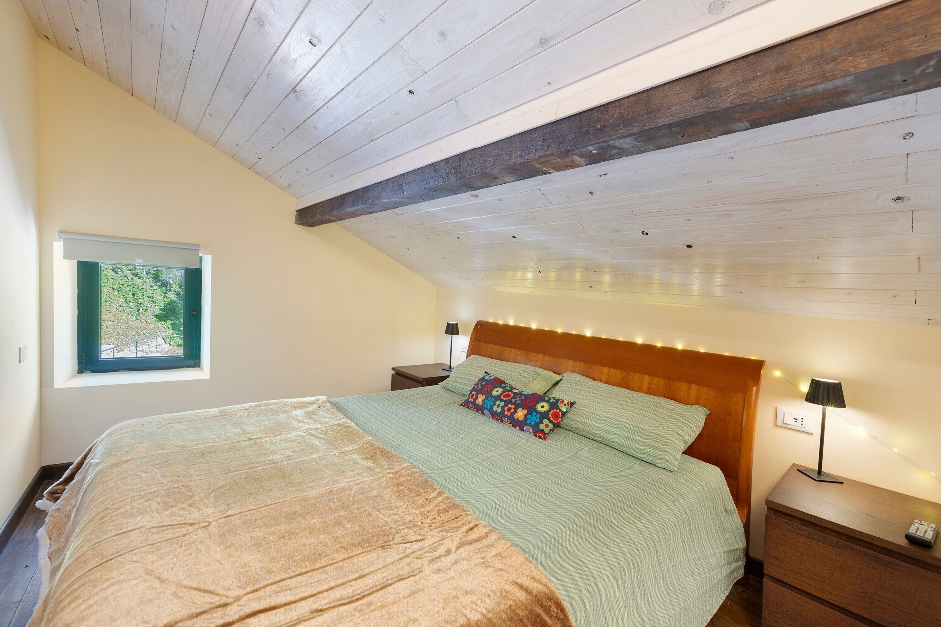 Een slaapkamer met een houten plafond, een bed met een groenbeige dekbedovertrek, een raam met uitzicht op groen en nachtkastlampen.